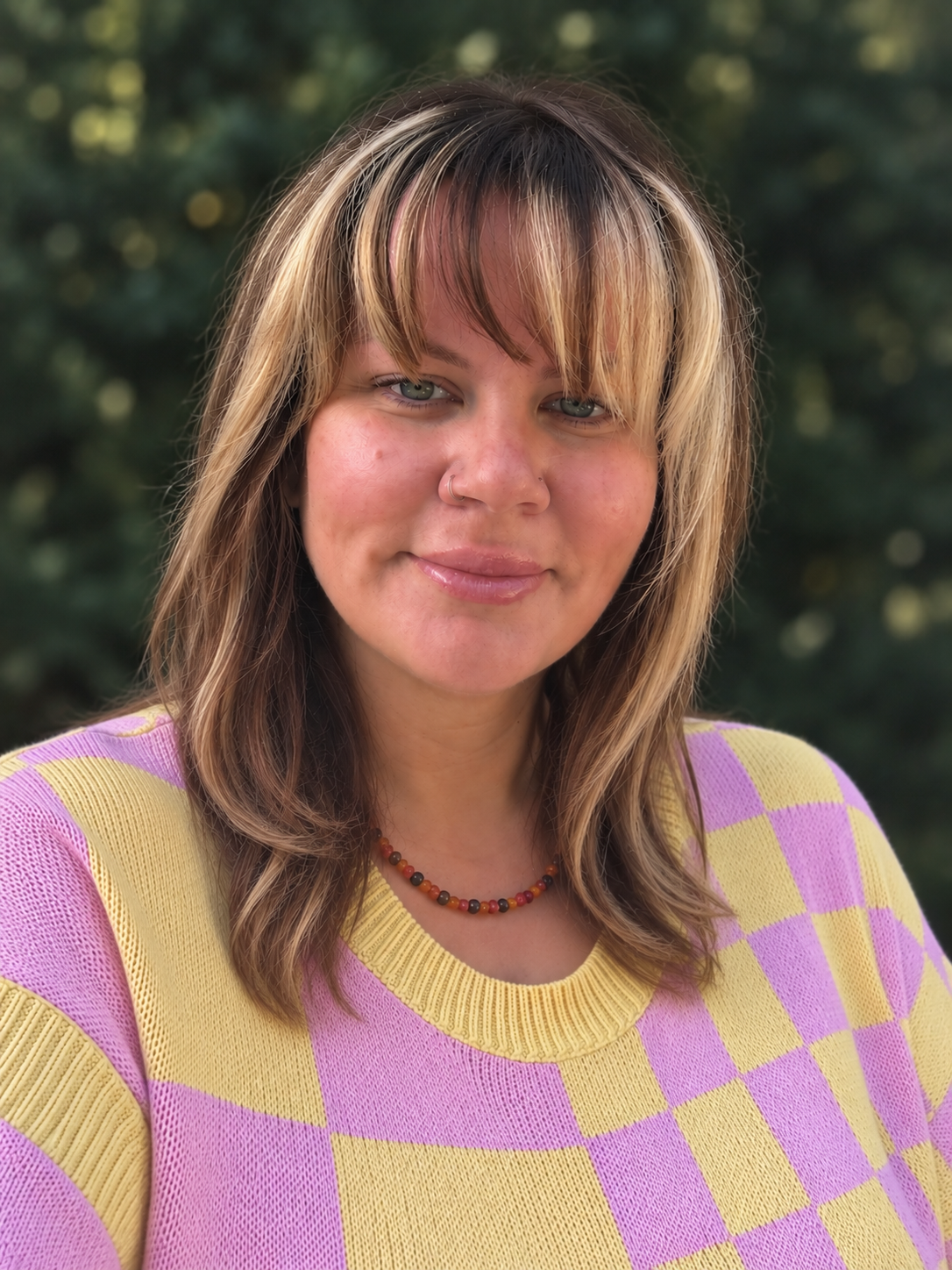 Portrait of Cassandra DeBolt Arnett smiling outdoors, wearing a pink and yellow checkered sweater